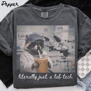 以下が含まれることがあります： ラボコート、サングラス、'lab is my jam'と書かれた野球帽を着用したアライグマのグラフィックがプリントされたチャコールグレーのTシャツ。アライグマはストロー付きのプラスチックカップを持っています。グラフィックの下には'literally just a lab tech'というテキストがあります。