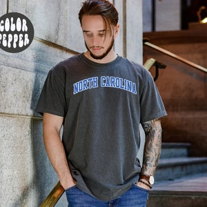 May include: Dark gray t-shirt with the text "NORTH CAROLINA" in blue arched lettering. The shirt has a vintage, washed look. The image also includes a "COLOR PEPPER" logo in a white bubble font.