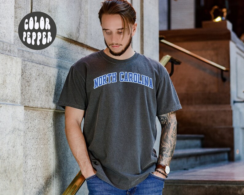 May include: Dark gray t-shirt with the text "NORTH CAROLINA" in blue arched lettering. The shirt has a vintage, washed look. The image also includes a "COLOR PEPPER" logo in a white bubble font.