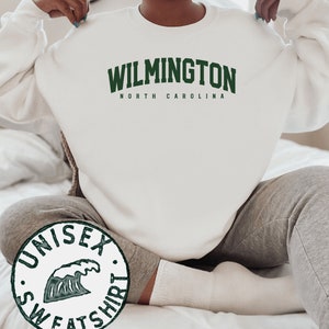 May include: White unisex sweatshirt with green text that reads "WILMINGTON NORTH CAROLINA". A green wave graphic is on a separate circular tag that reads "UNISEX SWEATSHIRT".