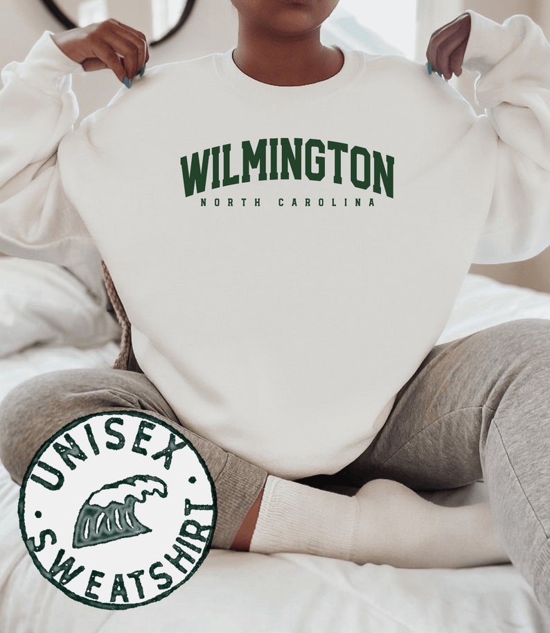 May include: White unisex sweatshirt with green text that reads "WILMINGTON NORTH CAROLINA". A green wave graphic is on a separate circular tag that reads "UNISEX SWEATSHIRT".