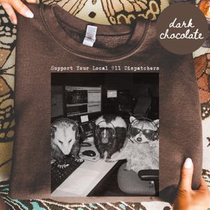 Puede incluir: Una sudadera de cuello redondo en color marrón chocolate oscuro con un estampado gráfico en blanco y negro de una zarigüeya, una mofeta y un mapache con auriculares en un escritorio. El texto superior dice Support Your Local 911 Dispatchers.