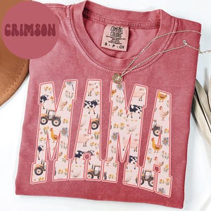 A pink t-shirt with the word "MAMA" printed on it, featuring various farm-themed illustrations such as tractors, animals, and flowers.