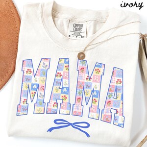 May include: An ivory-coloured t-shirt displaying the word "MAMA" in a patchwork floral design. The letters are outlined in blue and feature various pastel floral patterns. A blue bow is at the bottom. A gold necklace with a pendant is draped across the shirt.