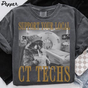 May include: A charcoal gray crew neck t-shirt featuring a graphic of a raccoon wearing a baseball cap and sunglasses while holding a drink. The text above and below the image reads SUPPORT YOUR LOCAL CT TECHS in a bold, gold-colored serif font.