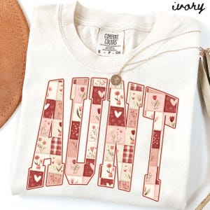 May include: Ivory-colored t-shirt with the word "AUNT" in a patchwork design. The letters are filled with floral and heart patterns in shades of red, pink, and cream. A gold necklace with a pendant rests on the shirt.