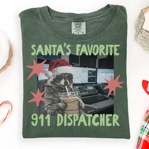 Pode incluir: Uma t-shirt verde musgo com um gráfico de um guaxinim usando um gorro de Pai Natal e óculos de sol, segurando uma bebida. O texto diz SANTA'S FAVORITE 911 DISPATCHER. O design inclui estrelas cor-de-rosa e luzes de Natal à volta do guaxinim.