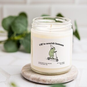 a candle labeled "Off to nourish humans" with a frog illustration, placed on a small stone or coaster.