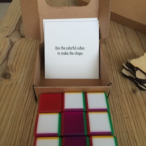 Puede incluir: Un conjunto de cubos coloridos dispuestos en una cuadrícula de 3x3, con lados rojos, blancos, verdes y morados. Los cubos están en una caja de cartón con asa. Una tarjeta dentro de la caja dice: "Usa los cubos de colores para hacer la forma."