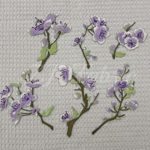 Pode incluir: Um conjunto de cinco emblemas bordados com flores de cerejeira em tom lilás sobre ramos castanhos com pequenas folhas verdes. Cada emblema apresenta costuras detalhadas com pétalas brancas, centros roxos e estames em roxo escuro.
