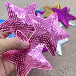 May include: A set of five sparkly star-shaped patches. The patches are made of sequins and come in a variety of colours, including pink, purple, gold, silver, and blue.