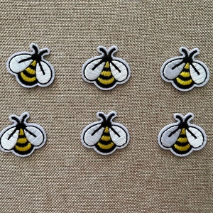 Może przedstawiać: Sześć haftowanych naszywek z motywem pszczół z żółto-czarnymi paskami i białymi skrzydłami. Naszywki znajdują się na beżowym tle z tkaniny.