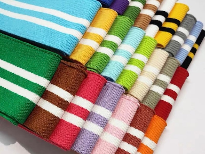 Op de afbeelding: Een assortiment van kleurrijke gebreide stofranden met witte strepen. De randen zijn gevouwen en gestapeld in een stapel. De randen zijn verkrijgbaar in kleuren zoals blauw, groen, geel, oranje, rood, bruin, paars, roze en grijs.