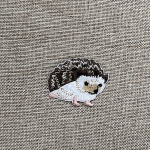 Puede incluir: Un erizo bordado de color marrón y blanco con una nariz marrón claro y patas rosadas. El erizo está sobre un fondo de arpillera marrón.