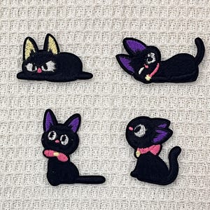 May include: Four black embroidered iron-on patches of cats with purple ears and pink collars. The cats are in different poses, including lying down, sitting up, and looking up.