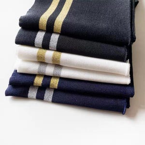 May include: A stack of six folded knitwear items in navy, black, and white. Each item features a contrasting stripe in either gold or silver. The items are neatly arranged, showcasing the different color combinations and textures.