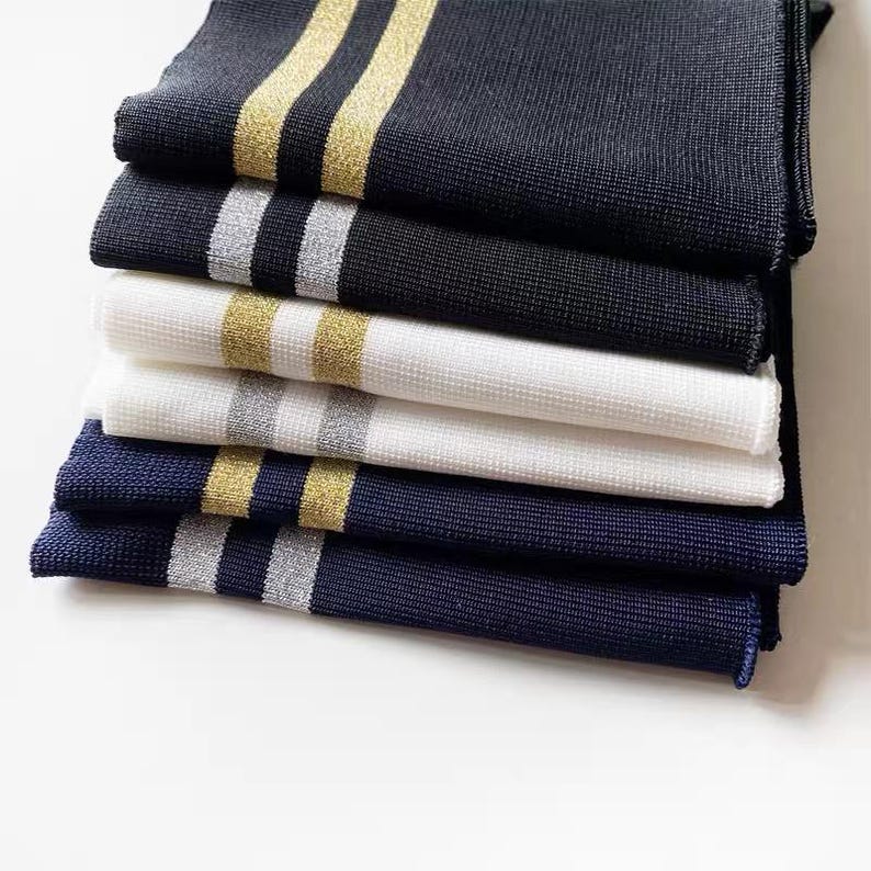 May include: A stack of six folded knitwear items in navy, black, and white. Each item features a contrasting stripe in either gold or silver. The items are neatly arranged, showcasing the different color combinations and textures.