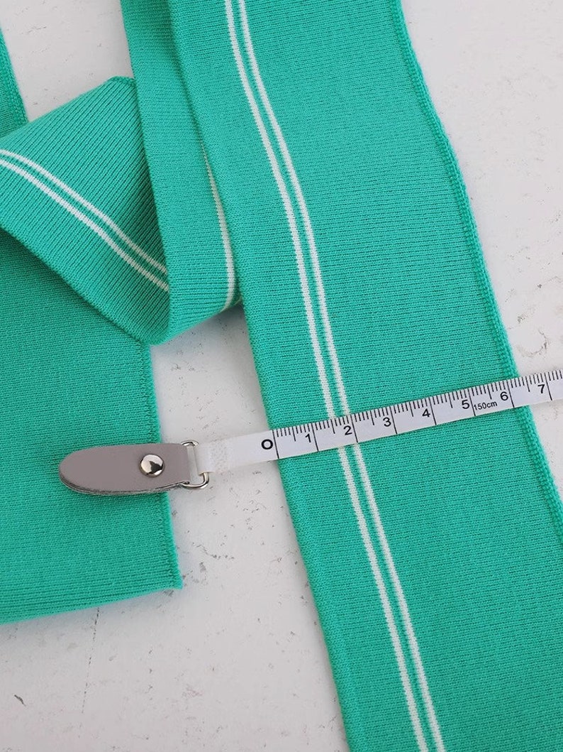 May include: A teal blue knit fabric with two white stripes running down the length of the fabric. A white measuring tape is laid across the fabric, showing 7 inches.