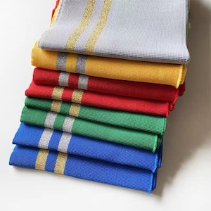 May include: A stack of colorful, folded fabric napkins. The napkins are in shades of gray, yellow, red, green, and blue. Each napkin has two metallic gold or silver stripes. The napkins are neatly stacked, showcasing the variety of colors and patterns.
