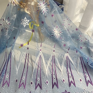 May include: Light blue tulle fabric with a white snowflake pattern and purple glitter accents. The fabric has a repeating pattern of white and purple stylized icicles.