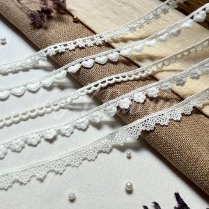 May include: A collection of five distinct white lace trims arranged diagonally. The trims feature various patterns including small floral motifs, scalloped edges, and heart-shaped designs, all crafted with intricate white thread detailing.