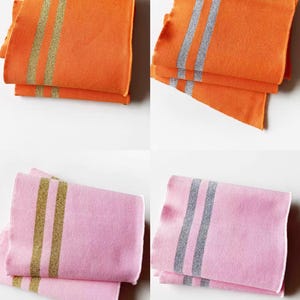May include: Four folded cloth napkins in two colors: orange and pink. The orange napkins have gold stripes, while the pink napkins have silver stripes. The napkins are stacked in pairs, showcasing the contrasting colors and designs.