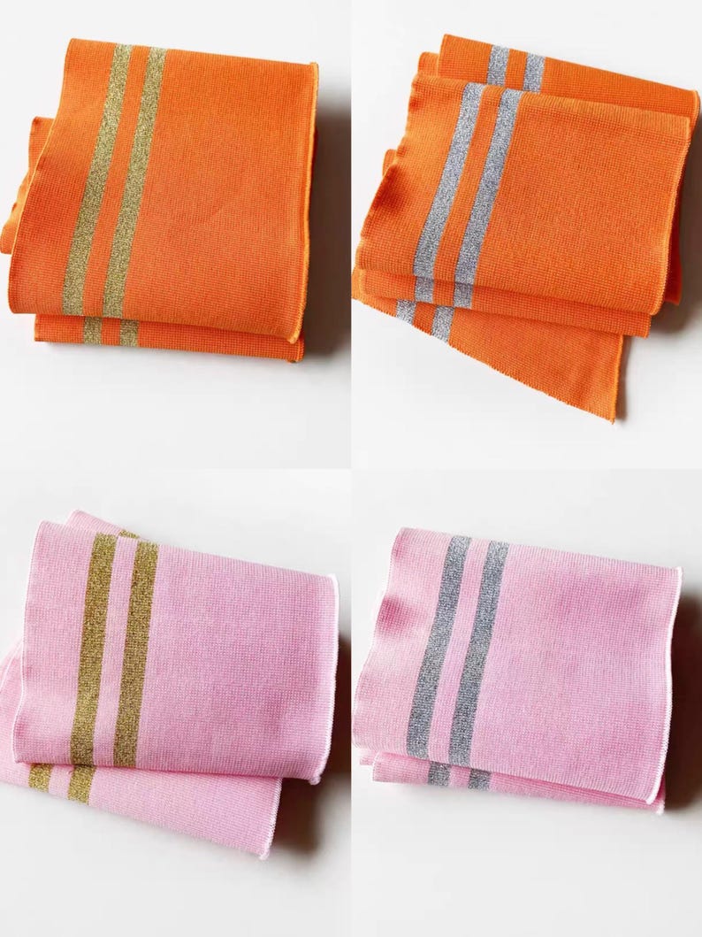 May include: Four folded cloth napkins in two colors: orange and pink. The orange napkins have gold stripes, while the pink napkins have silver stripes. The napkins are stacked in pairs, showcasing the contrasting colors and designs.