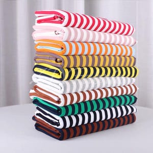 Può includere: Una pila verticale di pezzi di tessuto piegati, ognuno caratterizzato da un distinto motivo a righe orizzontali. La collezione include varie combinazioni di colori come righe rosse e bianche, rosa e bianche, arancioni e bianche, gialle e nere, grigie e bianche, marroni e bianche, verdi e nere, e blu navy e bianche.