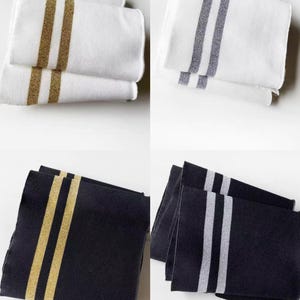 May include: Four sets of decorative towels. Two sets are white with gold or silver stripes. The other two sets are black with gold or silver stripes. The towels are folded and stacked, showcasing the striped design.