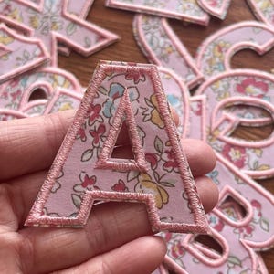 Puede incluir: Una letra A de tela rosa con un patrón floral y costuras rosas. La letra se sostiene en una mano.