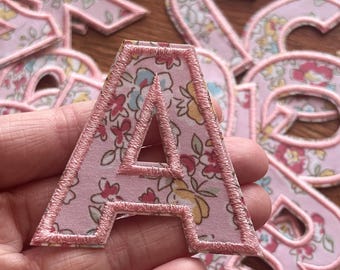May include: A pink fabric letter A with a floral pattern and pink stitching. The letter is held in a hand.