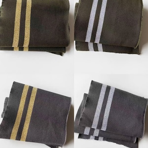 May include: Four folded scarves in shades of olive green and charcoal gray. Two scarves have gold stripes, and two have silver stripes. The scarves are made of a textured fabric and are neatly folded.