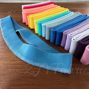 May include: A collection of colorful, ribbed elastic bands in a rainbow of hues, including pink, yellow, blue, and purple. A single, light blue elastic band is laid out in the foreground. The bands are neatly stacked, showcasing their texture and color variations.