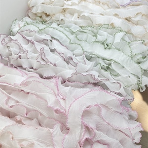 Puede incluir: Primer plano de varias filas de ribete blanco con volantes y bordes de diferentes colores. El ribete está dispuesto en forma de cascada, con la fila superior de color morado, la siguiente fila de color beige, la siguiente fila de color verde y la fila inferior de color rosa.