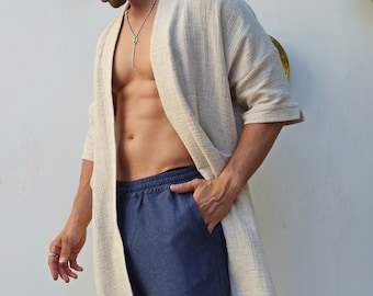 May include: A man wearing a light beige linen robe over a pair of dark blue pants. He is wearing black sandals and sunglasses.
