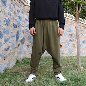 May include: A man wearing a black long-sleeved shirt with a button-up collar and olive green pants. He is standing in front of a stone wall with a small tree in the background.