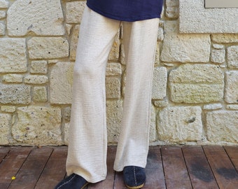 May include: A person wearing a navy blue long-sleeved shirt with a silver chain necklace and beige trousers. The shirt has a button-up collar and a pocket on the left side. The person is standing in front of a stone wall with a window.