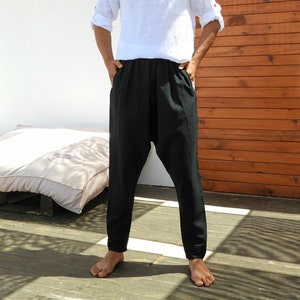 May include: A man wearing a white linen shirt with a button-up collar and black pants. He is standing in front of a wooden wall.