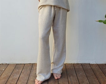 May include: A person wearing a light beige sleeveless top and matching trousers. The top has a V-neck and the trousers have a drawstring waist. The person is standing on a wooden floor.