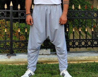 May include: A man wearing a white t-shirt and gray harem pants. He is standing in front of a black metal fence with gold accents.