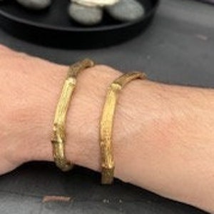 Puede incluir: Dos pulseras de metal dorado con una superficie texturizada ligeramente rugosa. Las pulseras están abiertas y tienen un diseño simple y minimalista.