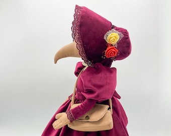 May include: A burgundy velvet doll with a long, pointed nose, wearing a burgundy velvet bonnet with lace trim and a small bouquet of fabric flowers. The doll is holding a brown handbag and has a lace trim on the bottom of the dress.