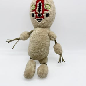 Puede incluir: Un juguete de peluche beige con una cara roja y negra y ojos verdes. El juguete tiene brazos y piernas largos con ap&eacute;ndices de cuerda.