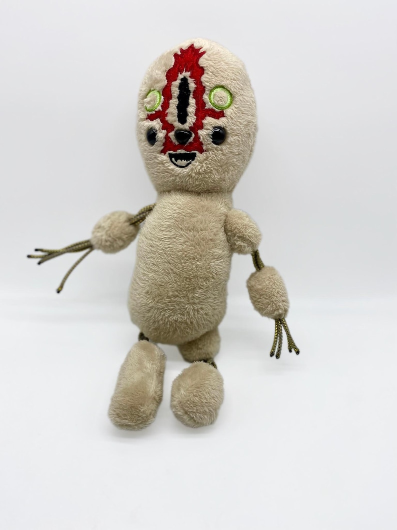 Puede incluir: Un juguete de peluche beige con una cara roja y negra y ojos verdes. El juguete tiene brazos y piernas largos con ap&eacute;ndices de cuerda.
