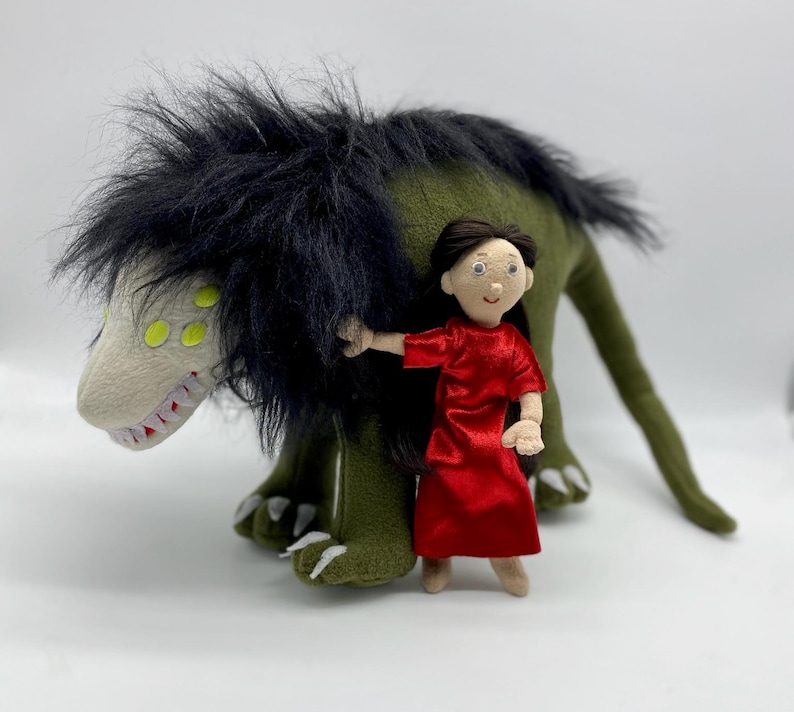 May include: A plush green monster with black fur and yellow eyes, holding a small doll with brown hair wearing a red dress.
