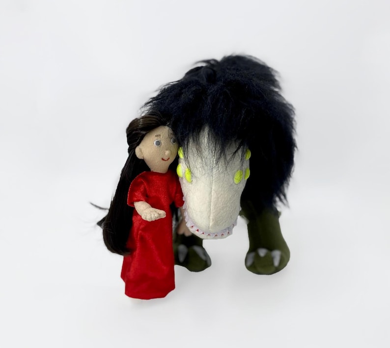 May include: A plush toy with black fur and green feet, with a white face and yellow eyes, is being hugged by a doll with long black hair wearing a red dress.