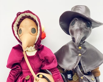 May include: Two plush dolls dressed in vintage plague doctor costumes. The female doll wears a burgundy dress with a lace trim and a beige satchel. The male doll wears a brown leather coat with a brown satchel and a black hat.