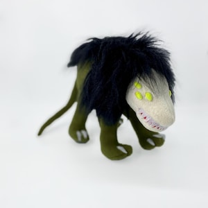 May include: Green and black plush toy with a long tail and four legs. The toy has a white face with yellow eyes and sharp teeth.