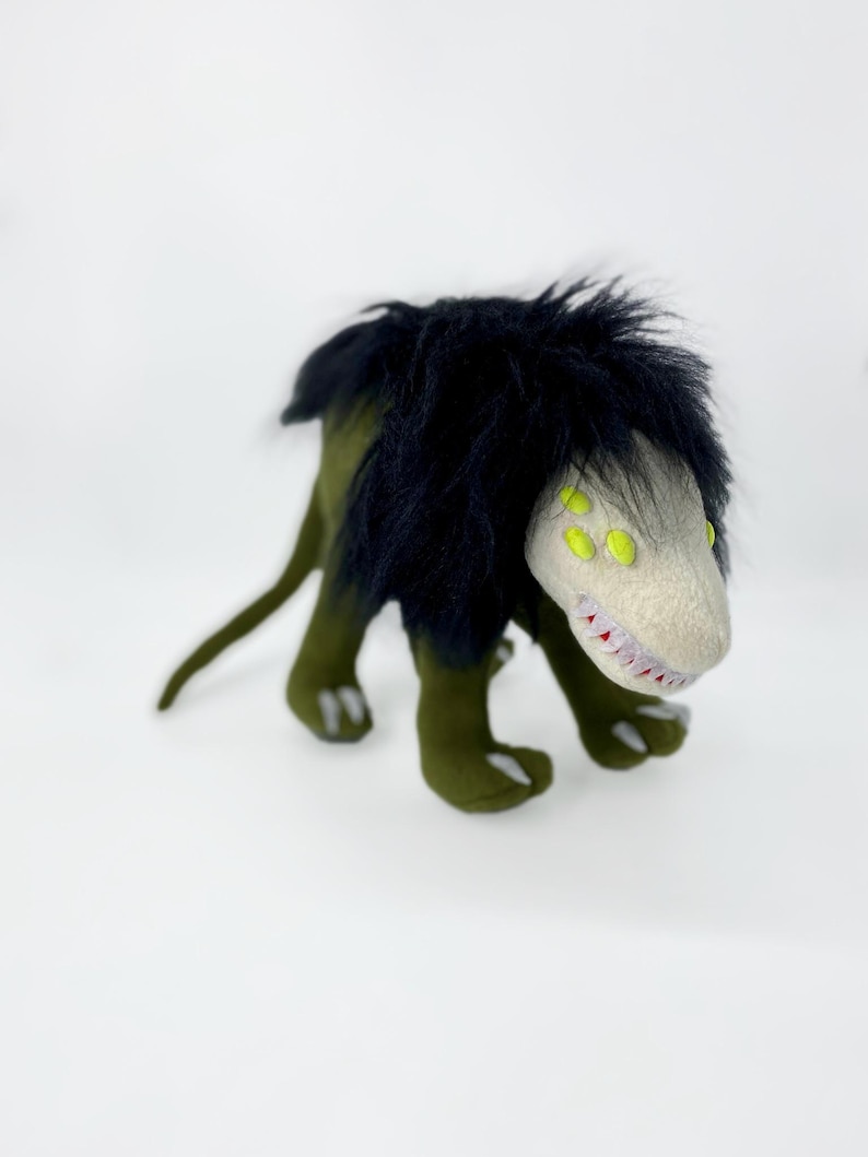 May include: Green and black plush toy with a long tail and four legs. The toy has a white face with yellow eyes and sharp teeth.