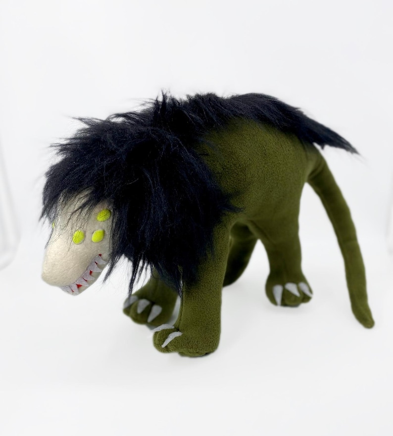 May include: Green plush monster toy with black fur, yellow eyes, and sharp teeth. The monster has a long tail and is standing on a white surface.